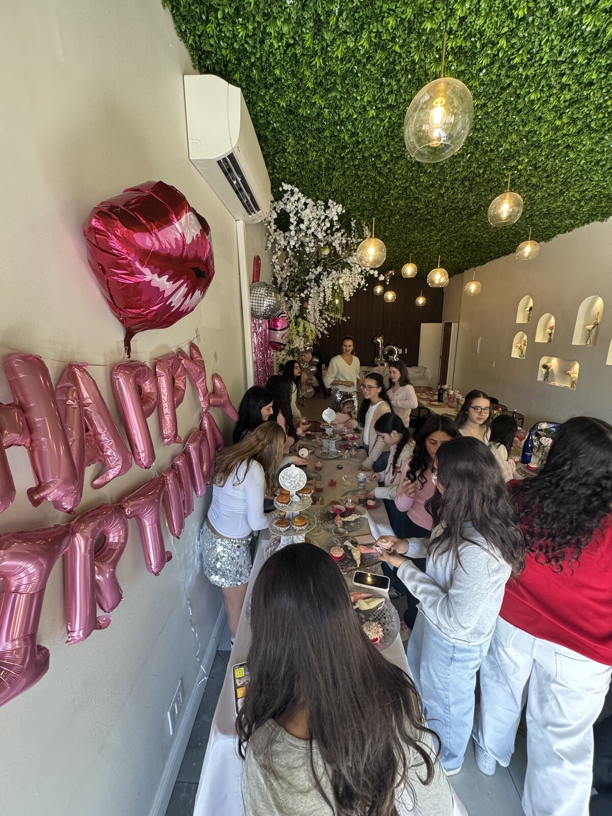 Birthday party guests around a long table under a greenery ceiling with pink Happy Birthday balloons